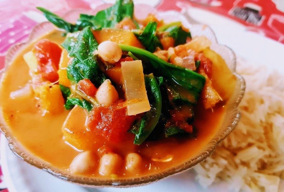 A glass bowl of vibrant vegetables, organge pumpkin, green spinach, and chickpeas. On a white plate with cooked rice
