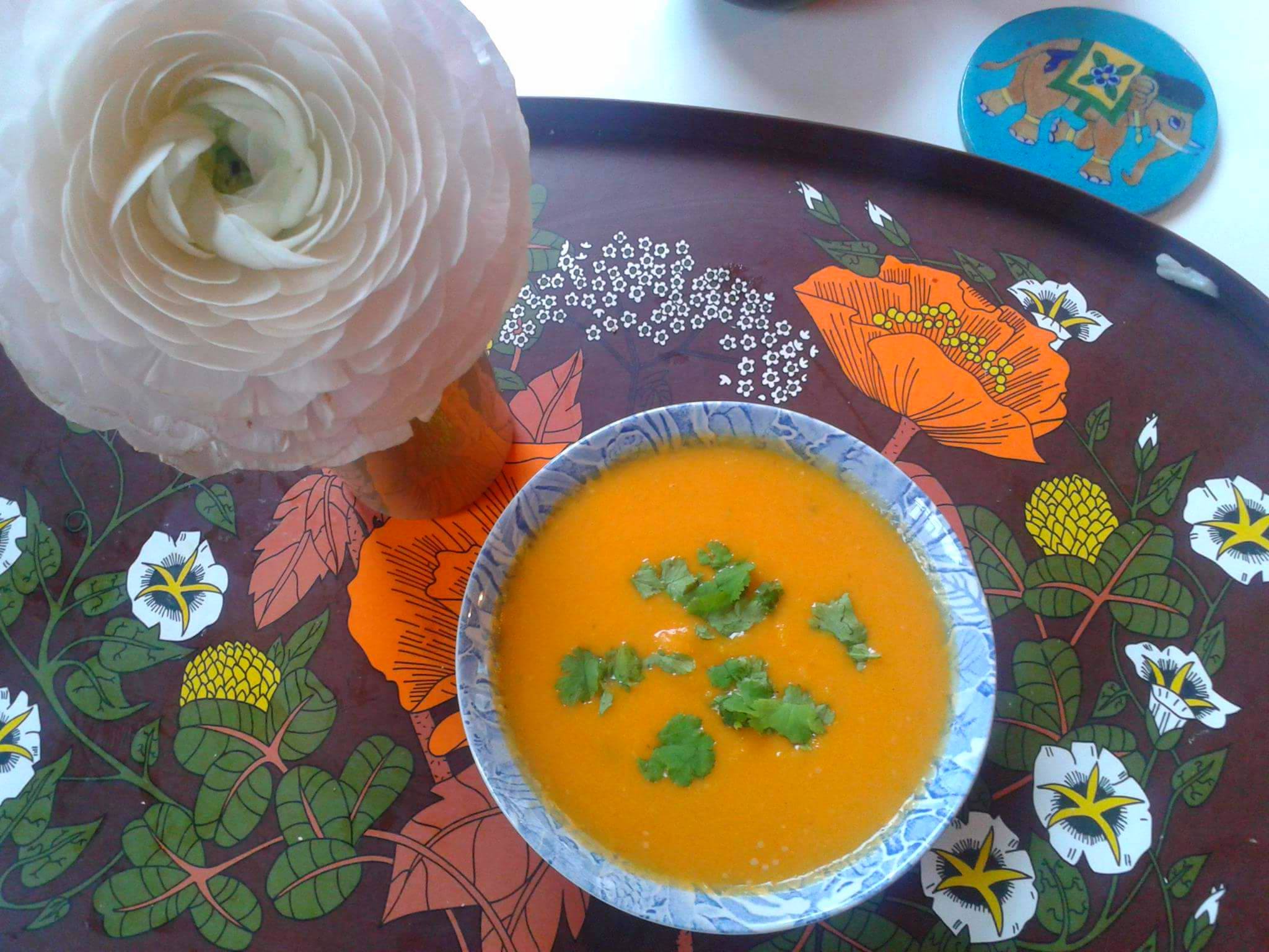 A bowl of vibrant orange soup on a 1970s vintage tray with orange flowers printed on it and a large pink rose stem in a vase